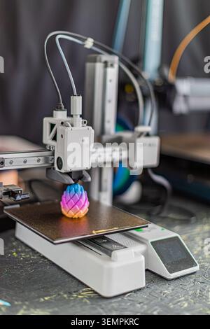 Multiaxis 3D printer with metallic frame, white extruder head depositing material onto dark build plate, showcasing gradient color scheme from blue to Stock Photo