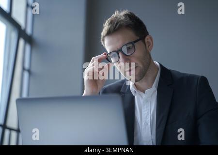 Businessman in suit adjusting glasses and concentrating on laptop screen while working in modern office Stock Photo