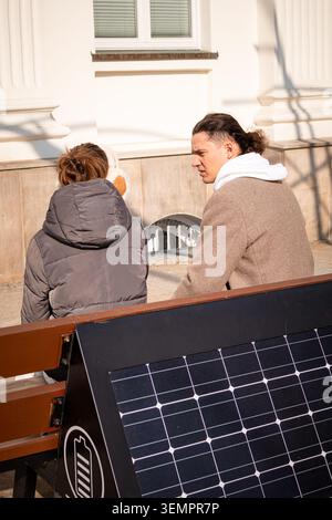 Father and daughter sitting on a smart solar-powered bench, talking and spending time together while charging a smartphone. Concept of family bonding, Stock Photo