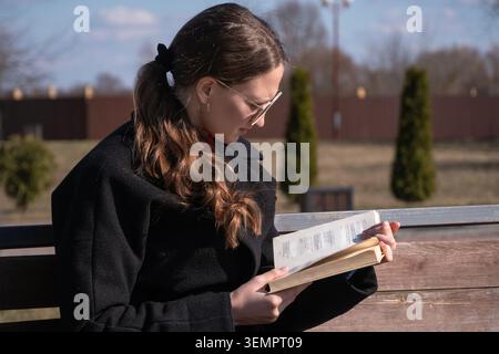 Young woman in sunglasses and a black coat sits on a wooden park bench reading a book, enjoying peaceful outdoor leisure and sunshine in an urban park Stock Photo