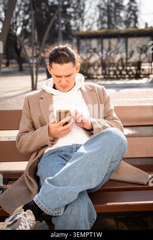Middle aged man sitting on bench and using smartphone outdoors in sunny city environment. Mobile communication, remote work, digital lifestyle and mod Stock Photo