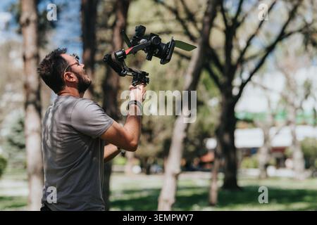 Man operating a handheld camera gimbal rig with propellers outdoors in a sunlit park Stock Photo
