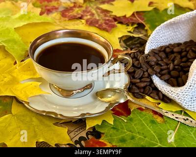 Black coffee in a vintage cup with a silver gold plated spoon on a saucer and natural coffee beans on burlap against a backdrop of autumn maple leaves Stock Photo