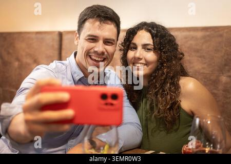 Smiling young couple taking a selfie with a smartphone while enjoying dinner at a restaurant. Modern lifestyle, social media sharing, leisure time, da Stock Photo