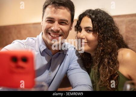 Smiling young couple taking a selfie with a smartphone while enjoying dinner at a restaurant. Modern lifestyle, social media sharing, leisure time, da Stock Photo