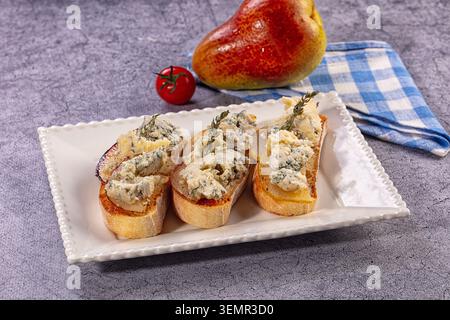 Three slices of toasted bread topped with pear slices crumbled blue cheese, garnished with thyme. Served on a white rectangular plate with a whole pea Stock Photo