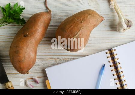 Two sweet potatoes on a light wooden surface next to an open notebook with and a blue pen. The atmosphere of the photo conveys the process of planning Stock Photo