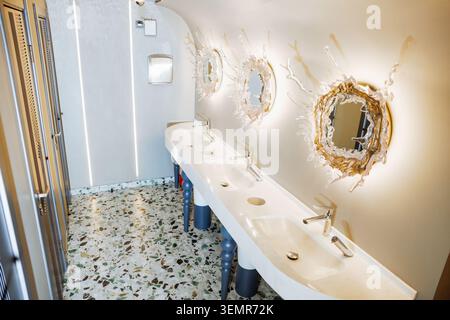 Wide perspective of a high-end public restroom featuring a long white vanity with three sinks, unique blue sculptural legs, and organic backlit glass Stock Photo