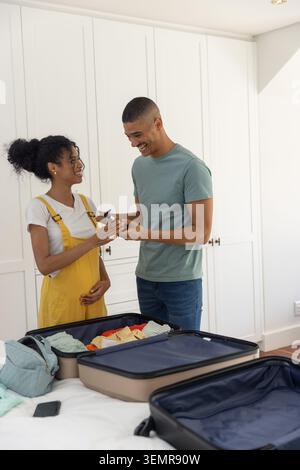 African American couple packing suitcases on bed in bedroom, woman in yellow overalls, phone on bed Stock Photo