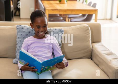 African American child sitting on sofa at home, reading bright-blue kids' book by light-blue pillow Stock Photo