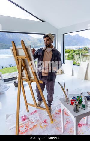 Male painter standing at wooden easel in bright studio, holding palette and brush, wearing beanie Stock Photo