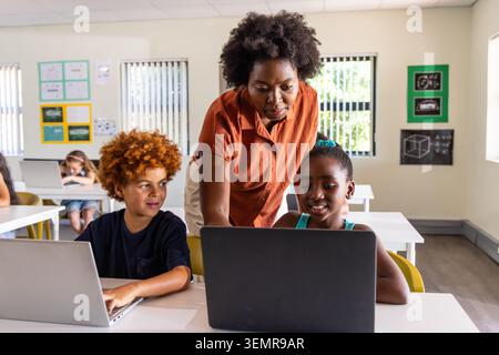 Diverse teacher in rust shirt leaning over desk aiding two children using two laptops in classroom Stock Photo