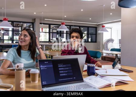 Diverse college students studying at communal table, using laptops, notebooks, travel mug Stock Photo
