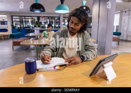 Indian man and Asian man working, writing in notebook at co-working lounge near tablet and tumbler Stock Photo