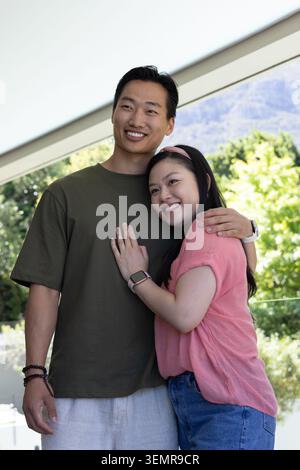 Asian couple standing on covered patio under slanted roof hugging, smiling, showing smartwatches Stock Photo