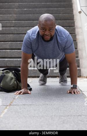 Adult African man doing push-ups on slabs at base of stairs, wearing heather tee, smartwatch Stock Photo