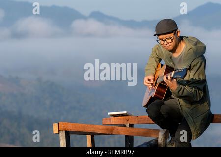 A young tourist plays an acoustic guitar on a wooden balcony at a mountain summit during the morning. Stock Photo