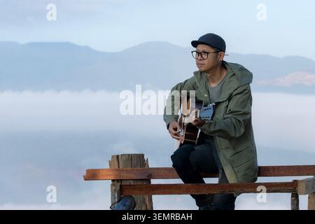 A young tourist plays an acoustic guitar on a wooden balcony at a mountain summit during the morning. Stock Photo