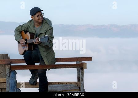 A young tourist plays an acoustic guitar on a wooden balcony at a mountain summit during the morning. Stock Photo