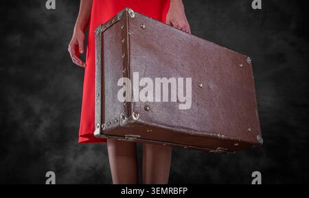 A woman holds an old brown vintage suitcase against a dark textured background. Stock Photo