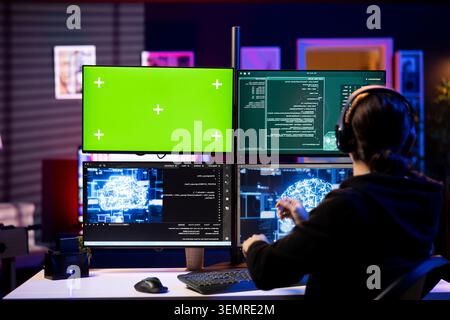 Hacker listening music using AI on green screen PC to steal credit card numbers and infiltrate banking systems, with cybercriminal wearing headphones running malicious code on chroma key computer. Stock Photo