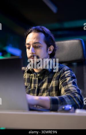 Serious millennial man using laptop sitting at the table, focused casual guy communicating online, writing emails, distantly working or studying on his portable computer in his apartment. Stock Photo