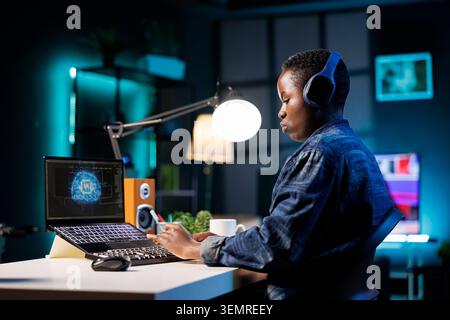 Side-view of black woman working at a desk, programming software and managing a futuristic neural network interface on her laptop, female developer monitoring database system on personal computer. Stock Photo