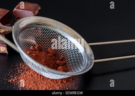 Metal sieve with cocoa powder and dark chocolate pieces on a black background, moody baking still life with rich texture and elegant copy space Stock Photo