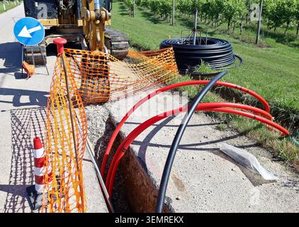 Road works for the laying of optical fibers under the roadway with black and red pipes. Around the excavation  safety signs, excavator and fence Stock Photo