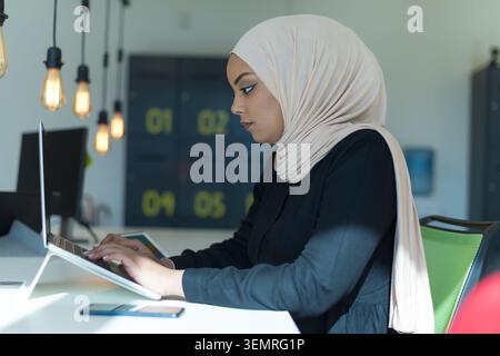 Side-view portrait of a young Muslim woman in a beige hijab and black top focused entirely on her work, typing on a laptop positioned on an ergonomic Stock Photo