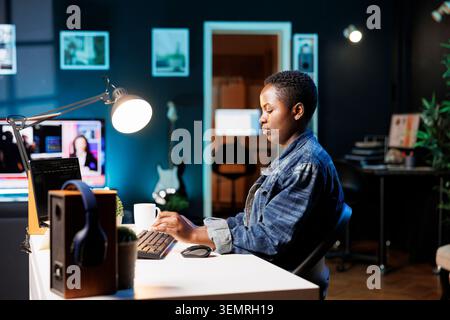 African American woman working at a desk with a laptop in her apartment, female self-employed individual engrossed in her personal computer, machine learning and monitoring database Stock Photo
