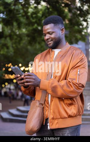 Adult African man standing in plaza at evening using smartphone wearing bomber jacket crossbody bag Stock Photo