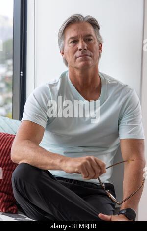 Man sitting on couch near tall window holding eyeglasses, wearing mint-green T-shirt and smartwatch Stock Photo
