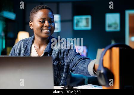 Young African American freelancer works from home on laptop while sticking a sticky note to electronic speaker, while using her personal computer, smiling black woman puts a post-it note on device Stock Photo