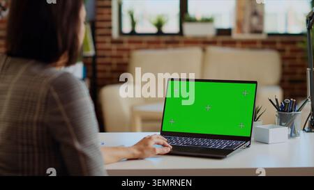 Freelancer checking emails on green screen laptop during work from home at computer desk. Self employed woman in cozy bright apartment doing job tasks, reading business messages on mockup device. Stock Photo