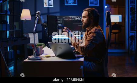 Developer writing lines of code on multimonitor PC setup during remote job shift. IT expert doing multitasking in neon lit home office, listening music, drinking coffee. Stock Photo