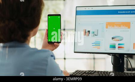 Manager holds phone with green screen in business agency workplace. Businesswoman working with isolated mockup template while showing blank chroma key display, startup job tasks. Stock Photo