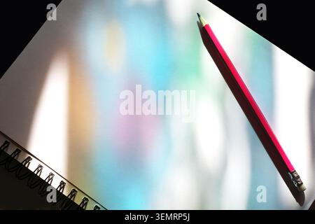 A red pencil rests on a white drawing block with a spiral binding on the left. The surface of the paper displays light effects in shades of blue, gree Stock Photo