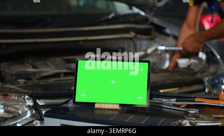 Tablet placed on working bench in busy garage next to professional tools while technician mends vehicle. Mockup device in repair shop with meticulous specialist working in background. Stock Photo