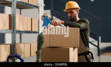 Manager wearing protective gear in warehouse teaching African American trainee how to seal cardboard box packages. Stock Photo