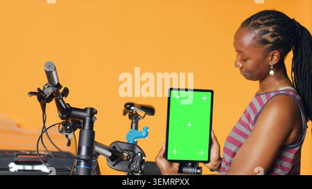 Woman holds green screen tablet, using it to advertise bike repair shop, empty placeholder for company logo, studio background. Mechanic promoting bicycle fixing service with mockup device. Stock Photo
