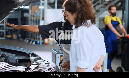 Helpful engineer assisting customer with car maintenance in auto repair shop. Licensed expert in garage looking over car components with woman, servicing her vehicle during annual checkup. Stock Photo