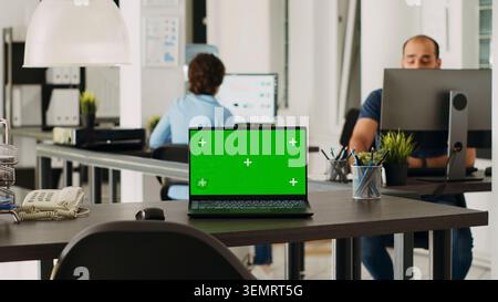 Laptop with green screen display on desk in modern startup agency office, wireless computer running isolated mockup copy space, with empty workstation with chroma key blank device. Stock Photo