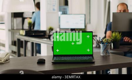 Green screen laptop in coworking space placed on empty office desk, open floor plan business workplace with isolated display, with portable computer running blank chroma key layout on software. Stock Photo