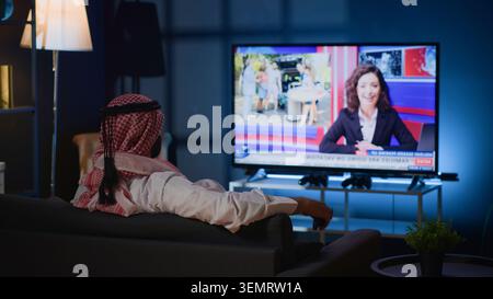 Arabic man lounged on cozy sofa, watching news broadcast while relaxing in apartment after hard day at work, with Middle Eastern person chilling on couch, looking at TV channel showing current events. Stock Photo