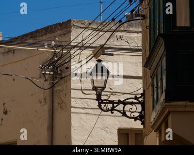 In Victoria, Malta, a street corner shows power lines and a vintage lamp against a pale wall under a blue sky. Stock Photo