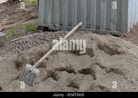 Shovel rests in gravel near a metal container. Sunlight highlights the rough texture of the sand. Background shows fencing and greenery beyond Stock Photo