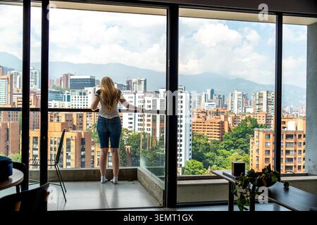 A woman stands at a glass railing inside a modern apartment, looking out over a dense urban landscape with tall buildings, green trees, and distant mo Stock Photo