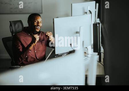 A man shows excitement in an office setting, sitting in front of several computer screens. He raises his hands and smiles, indicating he achieved some Stock Photo
