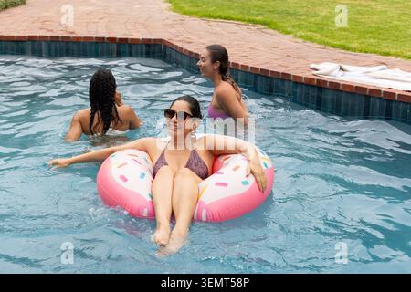 Diverse female friends relaxing in backyard pool in bikinis on pink donut float wearing sunglasses Stock Photo